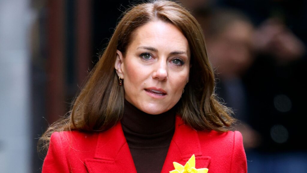 Princess Kate Pushed Back on One Aspect of Royal Family Life Kate Middleton wears a red coat with a black turtleneck sweater and a yellow daffodil on her lapel, she has long brown hair