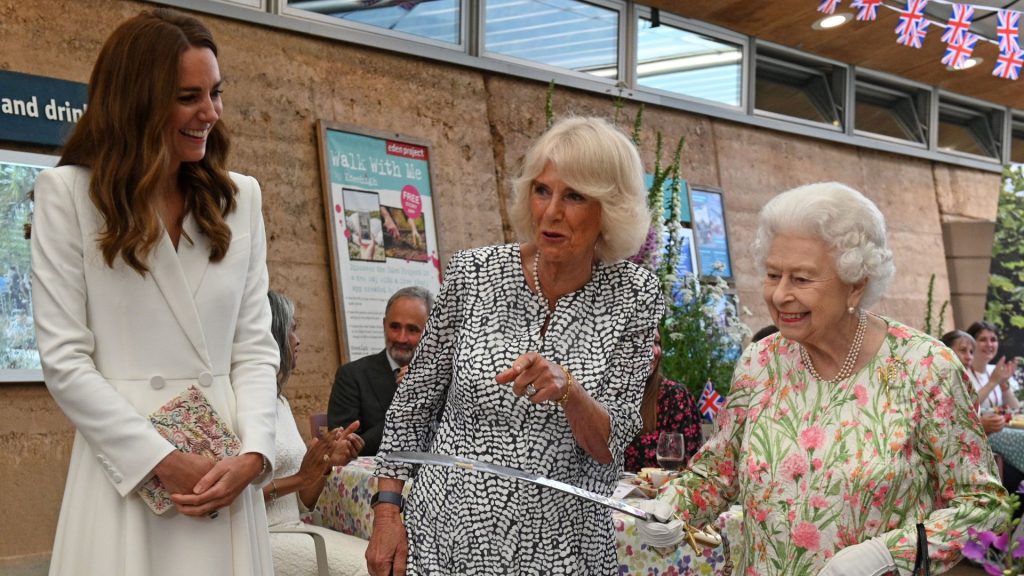 Princess Kate wears a white coat dress and has long brown hair, she laughs as Queen Camilla, wearing a black and white patterned dress, points and Queen Elizabeth holds a large knife