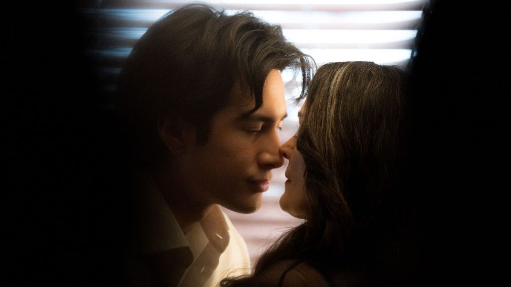 Hudson Williams and Carrie-Anne Moss stand closely together in front of window blinds, seen through shadows, in the Crave tv show 'Yaga.'