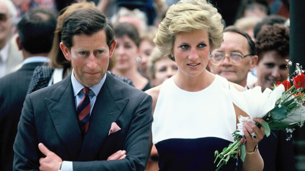 King Charles crosses his arms and wears a suit and striped tie while Princess Diana wears a dress with a white top and a black bodice while holding roses